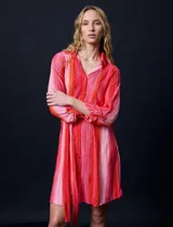 Vestido camisero midi de satén plisado con estampado a rayas verticales en tonos rosa y naranja. Tiene cuello camisero, cierre frontal con botones, mangas largas con puños abotonados y cinturón de lazo.