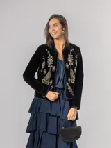 Chaqueta de terciopelo negro con bordados florales y ornamentales en tonos dorados, rosados y azules. Presenta un corte recto, cuello redondo y cierre frontal abierto con detalles de costuras decorativas en los bordes.
