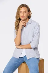 Camisa celeste y blanca a rayas verticales, de corte holgado, con cuello clásico, mangas 3/4 y bolsillo en el pecho.