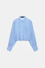 Camisa corta con estampado de cuadros vichy en tonos azul y blanco, cuello solapa, manga larga con puños anchos abotonados y bolsillo delantero de parche. Presenta bajo abullonado con elástico interior y cierre frontal de botones.