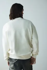 Buzo de tejido de algodón color crema, corte oversized y cuello redondo. Presenta un pequeño bordado central de Mickey Mouse vestido con ropa deportiva y sosteniendo una raqueta de tenis.