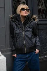 Campera negra de estilo urbano con diseño acolchado, cierre frontal dorado y cuello alto con textura. Incluye capucha con borde de piel sintética y puños de tejido acanalado.