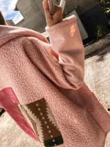 Campera tipo abrigo de textura borreguito en color rosa, con diseño de collage en la espalda que incorpora un parche de pana rojiza y un panel central con estampado geométrico étnico. Presenta capucha integrada y un detalle de etiqueta de cuero sintético en la parte posterior.
