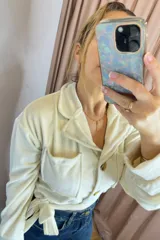 Campera tipo levita color crudo con textura efecto piel de durazno, cuello solapa, bolsillos frontales tipo cargo y cierre frontal con botones. Incluye un lazo ajustable en la cintura.