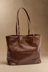 Bolso tipo shopper color marrón de cuero sintético con diseño trenzado, doble asa larga para el hombro y placa metálica frontal con grabado.