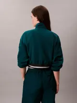 Campera tipo track Calvin Klein de tejido plano en color verde oscuro, de corte cropped y manga larga. Cuenta con cuello alto, cierre frontal de cremallera metálica, bolsillos laterales y elástico acanalado a rayas en los puños y el dobladillo. Presenta el logo de la marca bordado en el pecho.