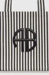 Bolso tipo shopper de diseño rectangular y amplio, confeccionado en tela con patrón de rayas verticales en color blanco y negro. Cuenta con dos asas superiores para llevar al hombro y detalle de aplique metálico con monograma en el centro.