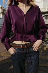 Camisa satinada de color violeta oscuro, con cuello solapa, escote en pico y manga larga. Presenta un detalle de cinturón fino ajustable a tono en la cintura y cierre frontal con botones.