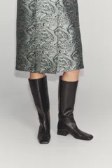 Bota de piel color marrón oscuro, de caña alta, con detalle de pespuntes en el cuerpo, tacón medio en bloque y punta cuadrada.