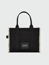 Bolso tipo tote de gran tamaño en color beige, confeccionado en lona resistente. Presenta el logo estampado en negro en el frente y detalles de cintas laterales con inscripciones. Cuenta con doble asa superior para llevar al hombro.