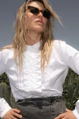 Blusa blanca de manga larga con cuello mao y pechera con pliegues verticales en forma de zigzag.