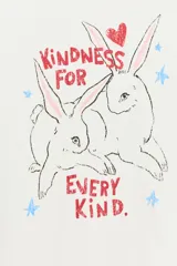 Camiseta blanca de algodón con cuello redondo, manga corta y calce regular. Presenta un estampado frontal ilustrado con conejos, corazones, estrellas y el texto "Kindness for every kind".