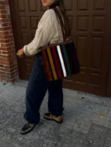 Bolso tipo tote de cuero con diseño de franjas verticales en tonos tierra, burdeos, verde y blanco. Cuenta con correas de hombro trenzadas y cierre superior.