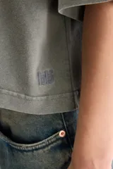 Remera cropped blanca de calce relajado, con cuello redondo, hombros caídos y mangas cortas. Presenta un pequeño detalle de etiqueta bordada en el lateral inferior.