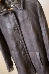 Abrigo de cuero marrón con cuello de piel sintética, cierre con botones y bolsillos laterales. Diseño con paneles geométricos y costuras a la vista.