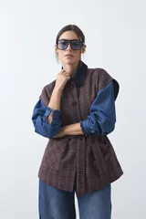 Chaleco de corte relajado con estampado a cuadros en tonos tierra, cuello camisero oscuro y cierre frontal con botones. Incluye un cinturón de la misma tela para ajustar a la cintura y bolsillos frontales.