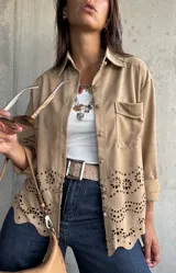 Sobrecamisa de gamuza color beige, con cuello camisero, cierre frontal con botones metálicos y bolsillo en el pecho. Presenta un diseño con calados florales y bordes ondulados en la parte inferior.