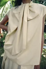 Camisa sin mangas confeccionada en algodón, con cuello redondo y detalle de lazada frontal. Presenta un diseño de corte holgado con bajo redondeado y cierre posterior mediante abertura y botones.