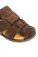 Sandalias tipo zueco de cuero gamuzado color marrón, con diseño de tiras cruzadas, puntera cerrada y una gran hebilla metálica dorada en el lateral. Cuenta con base de corcho y suela plana.