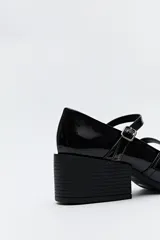 Zueco estilo Mary Jane color negro charol, con doble tira y hebillas metálicas. Presenta punta cuadrada y taco ancho de altura media.