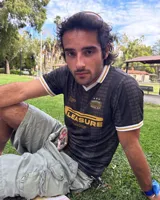 Camiseta de fútbol estilo retro de manga corta, color negro con diseño texturizado a rayas verticales, cuello en V blanco con detalle en mostaza y bordes de mangas a juego. Presenta estampados gráficos en el frente con la inscripción "PLEASURE" y emblemas deportivos.