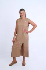 Vestido midi sin mangas de color tostado, con cuello redondo y tajo frontal. Presenta bordados artesanales de flores en tonos rosados y verdes distribuidos en el frente.