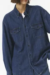 Camisa de denim azul oscuro, de corte semi-oversize con estilo vintage western. Presenta detalles de costuras en contraste, bolsillos exteriores y botones decorativos en el cuello.