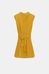 Vestido corto color mostaza con cuello fluido, manga sisa y hombreras marcadas. Presenta un detalle de lazada en la cintura y cierre de cremallera oculto en la espalda.