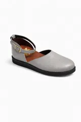 Zapato plano tipo bailarina o mary jane, color gris claro, con puntera redonda y tira de ajuste con hebilla en el tobillo. Suela negra.