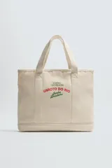 Bolso tipo shopper color crudo, fabricado en tejido de algodón con estampado de texto en verde y rojo. Tiene compartimento principal con cierre mediante imán, bolsillo interior con cierre y otro plano. Posee dos asas de mano.
