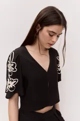 Blusa corta de color negro con escote en V, mangas cortas y bordados florales en contraste sobre las mangas.