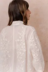 Camisa blanca de calce relajado con tejido semitransparente y bordados florales en relieve. Cuenta con cuello camisero, abotonadura frontal y mangas amplias.