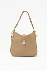 Cartera tipo hobo color beige, de diseño estructurado con cierre superior y una tira vertical con detalle de dije metálico circular en la parte frontal. Cuenta con asa corta trenzada y herrajes dorados.