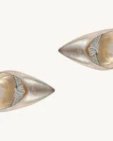 Zapato de tacón alto color dorado con pulsera al tobillo y detalle de moño plateado con brillos en la punta.