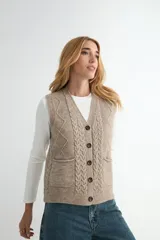 Chaleco de punto color beige con diseño de trenzas verticales y rombos en los laterales. Presenta cuello en V, cierre frontal con botones oscuros y dos bolsillos delanteros.
