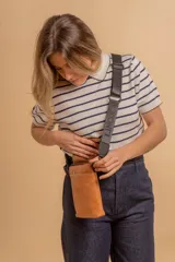 Phonebag pequeña color mostaza, confeccionada en cuero sintético, con solapa frontal y correa ajustable con logo de la marca.