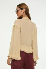 Blusa color tostado con escote en V profundo, volados en la parte inferior y en las mangas largas amplias, y un lazo frontal ajustable.