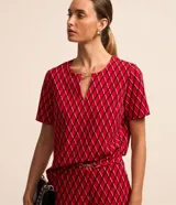 Blusa de crepé color rojo con estampado geométrico de rombos en negro y blanco. Tiene cuello redondo con abertura en forma de gota y detalle metálico, mangas cortas y corte recto.