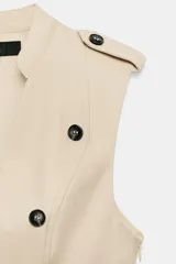 Vestido mini color beige sin mangas, con escote en V, doble botonadura frontal de botones oscuros y bajo con volante amplio. Presenta trabillas en los hombros y falsos bolsillos con solapa.