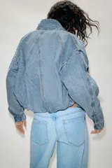 Cazadora bomber de denim con cuello subido, manga larga y cierre frontal con cremallera y botones a presión ocultos. Presenta costuras verticales decorativas y bajo con ajuste elástico.