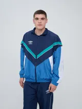 Campera deportiva Umbro modelo Beat, confeccionada en nylon ligero con diseño de bloques de color en blanco, azul marino y verde azulado. Presenta cierre frontal, cuello alto y elásticos en puños y cintura.