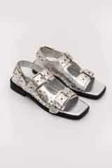 Sandalias negras de cuero con suela plana, dos tiras anchas con hebillas doradas y apliques metálicos.