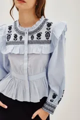 Camisa de algodón celeste con detalles de volados en los hombros y bordados en color negro en el pecho y las mangas. Presenta cierre frontal con botones y puños ajustados.