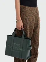 Bolso tipo tote de cuero negro, modelo The Leather Medium Tote Bag de Marc Jacobs. Presenta doble asa superior, correa ajustable para el hombro y el texto grabado "THE TOTE BAG MARC JACOBS" en el frente.