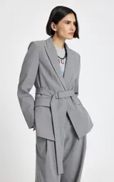 Blazer gris de sastrería con diseño entallado, cuello con solapas clásicas, hombros estructurados y mangas largas. Presenta bolsillos delanteros con solapa y un cinturón a juego con hebilla metálica para ajustar la cintura.