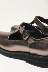 Zapato guillermina color bronce con acabado metalizado, con correa sobre el empeine con hebilla ajustable y suela de goma negra con plataforma.