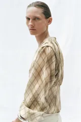 Camisa de manga larga con escote en V y cierre frontal con botones. Presenta un estampado de cuadros en tonos beige y marrón, con detalles de pliegues en el delantero y puños ajustados.