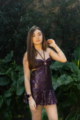 Vestido corto de lentejuelas color ciruela o morado oscuro, con escote corazón y detalle de argolla central que conecta con tirantes finos que atan al cuello. Presenta aberturas laterales (cut-outs) en la zona de la cintura y falda corta con vuelo.