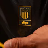 Camiseta de fútbol Puma de manga larga del Peñarol, edición aniversario 26, con diseño bicolor que combina mitades y rayas verticales en negro y amarillo, cuello tipo polo y detalles de sponsors en el frente.