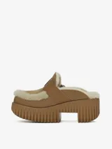 Zueco de plataforma alta confeccionado en cuero vacuno color tostado con forro de pelo de oveja. Presenta una correa con hebilla metálica ajustable en el empeine y suela de goma con diseño acanalado.